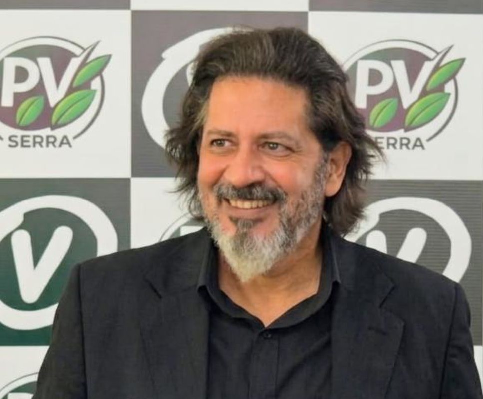 Primeiro plano e meio-corpo do jornalista e Secretário de Comunicação Lauro Nunes, em terno preto, sorrindo e olhando para a esquerda em pé diante de um painel de xadrez com os logotipos do PV Serra.
