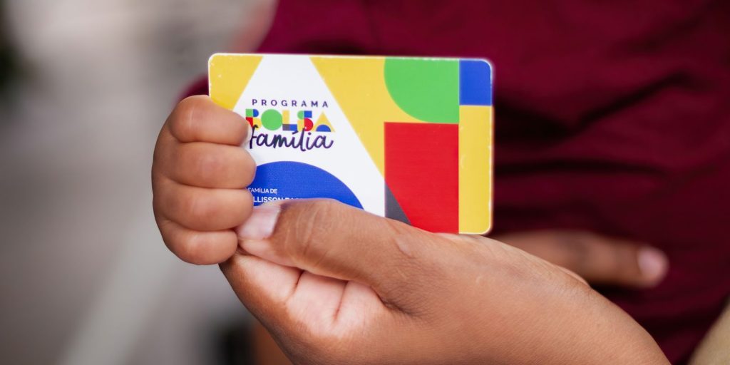 "Mãos de uma pessoa segurando o cartão oficial do programa Bolsa Família, com o logotipo colorido do governo federal. O foco está nos detalhes do cartão e o fundo está sutilmente desfocado, destacando o amparo social."