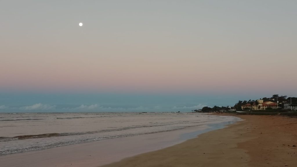 Paisagem da praia de Nova Almeida em Serra, ES, com o mar calmo, faixa de areia e a lua brilhando no céu durante o entardecer.