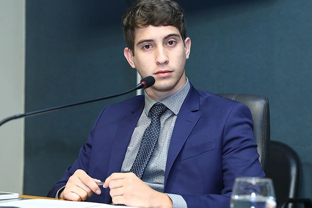 Deputado Lucas Polese Apresenta Requerimento para Instalação de CPI sobre a APEX no Espírito Santo Fotografia do deputado estadual Lucas Polese sentado à mesa de sessões, vestindo terno azul, camisa cinza e gravata escura, com um microfone à frente.