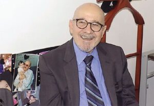 José Maria Ramos Gagno – 83 anos de um código de vida e liberdade Homem de meia-idade a idoso, careca, com óculos redondos e barba grisalha aparada, sentado e sorrindo. Ele veste um paletó escuro, camisa azul clara e gravata listrada em tons de azul e preto. Ao fundo, parede branca com quadros apoiados em uma superfície e um cabideiro de madeira.